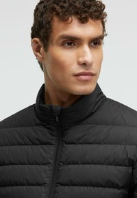 Black puffer jacket featuring a high collar and horizontal quilted design. Material is smooth with a lightweight, insulated appearance.