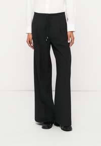 Mustad laiade sääreosadega püksid paelaga vöökohas, millel on lõhikud ja sileda tekstuuriga pind. Kandmiseks valge pika varrukaga särk.