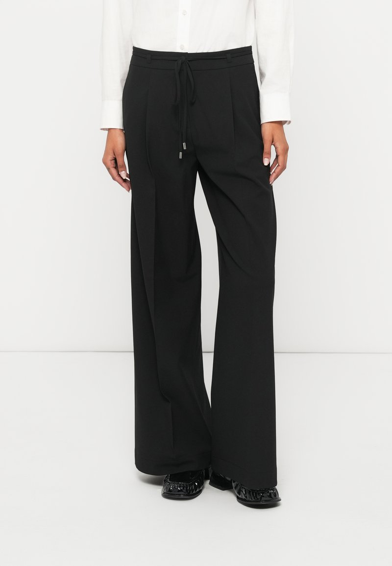 Mustad laiade sääreosadega püksid paelaga vöökohas, millel on lõhikud ja sileda tekstuuriga pind. Kandmiseks valge pika varrukaga särk.