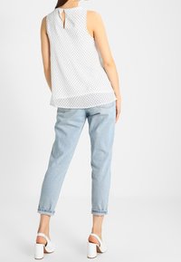 Witte mouwloze top met kleine zwarte stippen, achterkant met sleufdetail, gecombineerd met lichtblauwe denim jeans en witte blokhak sandalen.