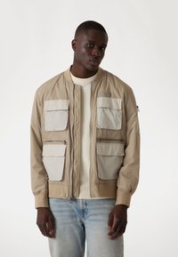 Veste bomber beige avec des poches beige clair, des poignets côtelés et des détails en zip. Portée par-dessus un pull crème avec un jean bleu clair.