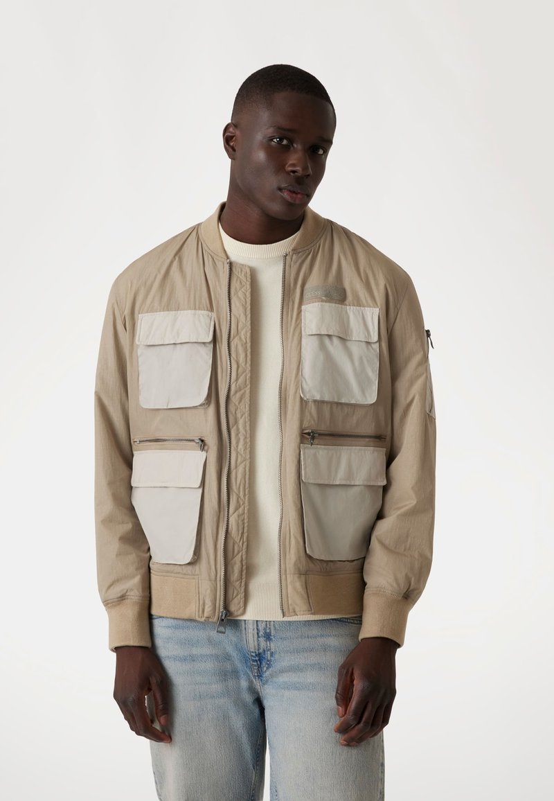 Veste bomber beige avec des poches beige clair, des poignets côtelés et des détails en zip. Portée par-dessus un pull crème avec un jean bleu clair.