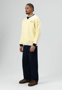 Gele trui met zwarte manchetten en kraag, met een logo op de borst. Gecombineerd met donkere spijkerbroek en bruine veterschoenen.