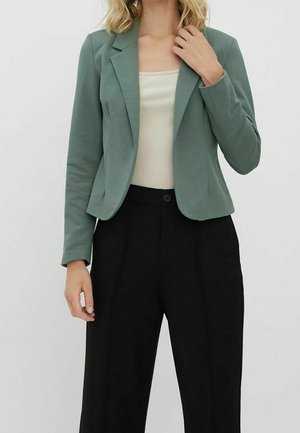 Femme portant un blazer vert sauge, un haut côtelé crème, et un pantalon noir à jambes larges taille haute sur un fond uni.
