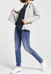 Světle šedý, otevřený kardigan přes námořnický vzorovaný top. Modré džíny s bílými postranními pruhy, doplněné bílými teniskami. Neformální outfit.