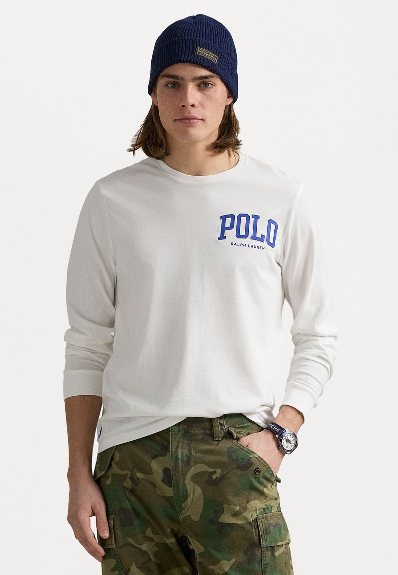 Λευκό πουκάμισο με μακριά μανίκια και μπλε λογότυπο "POLO", συνδυασμένο με πράσινα παντελόνια παραλλαγής. Διαθέτει ριμπ μανσέτες και ναυτικό πλεκτό σκουφάκι.