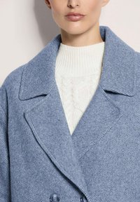 Lichtblauwe gestreepte wollen jas met een gestructureerde kraag, dubbelknopen ontwerp en blauwe knopen, gedragen over een crèmekleurige kabelbrei trui.