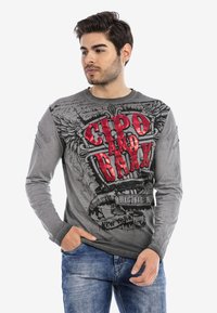 Graues Langarmshirt mit rotem und schwarzem Grafikdruck sowie Text. Verwaschenes Design mit Akzenten auf den Schultern. Kombiniert mit blauen Jeans.