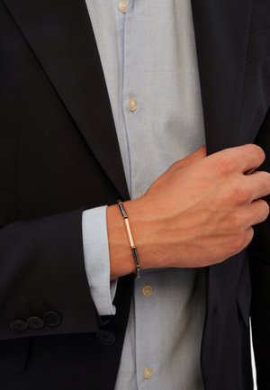 Bracciale in metallo con una barra dorata e accenti neri. Indossato su un polso, abbinato a un blazer scuro e una camicia azzurra chiara.