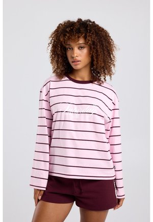 Vrouw met krullend haar draagt een roze en bordeaux gestreept shirt met lange mouwen en bordeaux korte broek, staand tegen een effen lichte achtergrond.