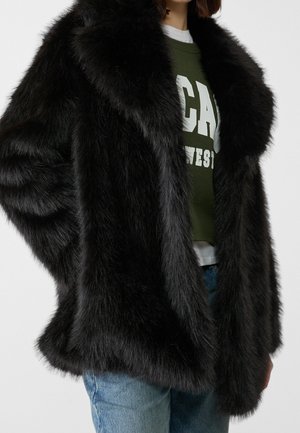 Vrouw draagt een zwarte nepbontjas over een groen sweatshirt met opdruk en blauwe spijkerbroek, zichtbaar van nek tot heupen tegen een effen achtergrond.