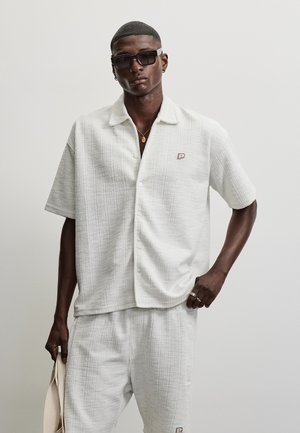 Bărbat purtând o cămașă albă texturată cu mâneci scurte și pantaloni, ochelari de soare negri, un colier de aur și inele, ținând un obiect din țesătură bej.
