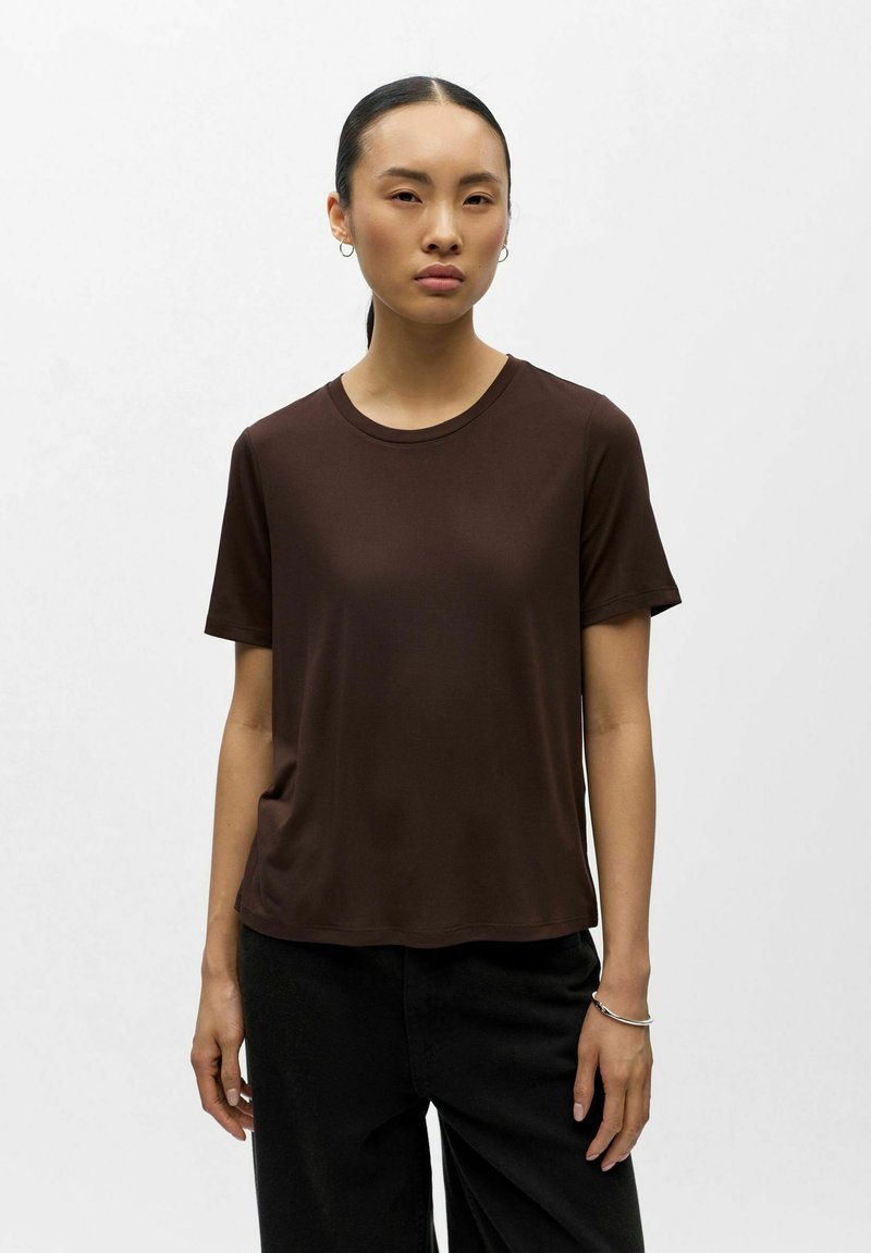 Femme aux cheveux noirs raides attachés, portant un t-shirt marron foncé à manches courtes et un pantalon noir, posant devant un fond uni.