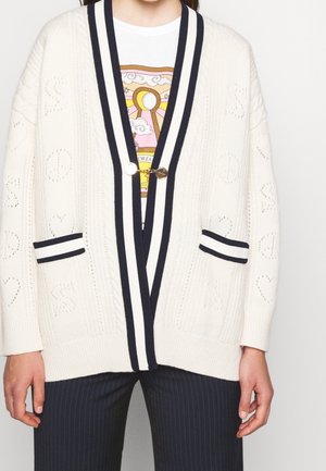 Cárdigan blanco de punto con ribete a rayas en blanco y negro, cierre con botones dorados, llevado sobre una camiseta gráfica y combinado con pantalones oscuros de rayas finas.
