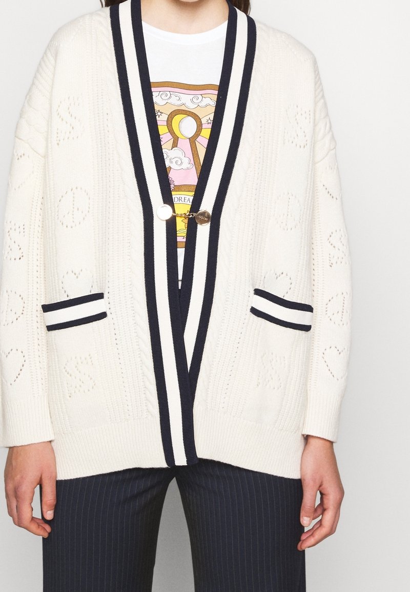 Cardigan blanc en maille avec bordure rayée noir et blanc, fermeture à boutons dorés, porté sur un t-shirt graphique et associé à un pantalon foncé à fines rayures.