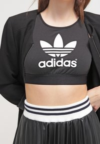 Crop top sportivo nero con logo Adidas bianco, abbinato a una gonna plissettata nera con una fascia in bianco e blu navy.
