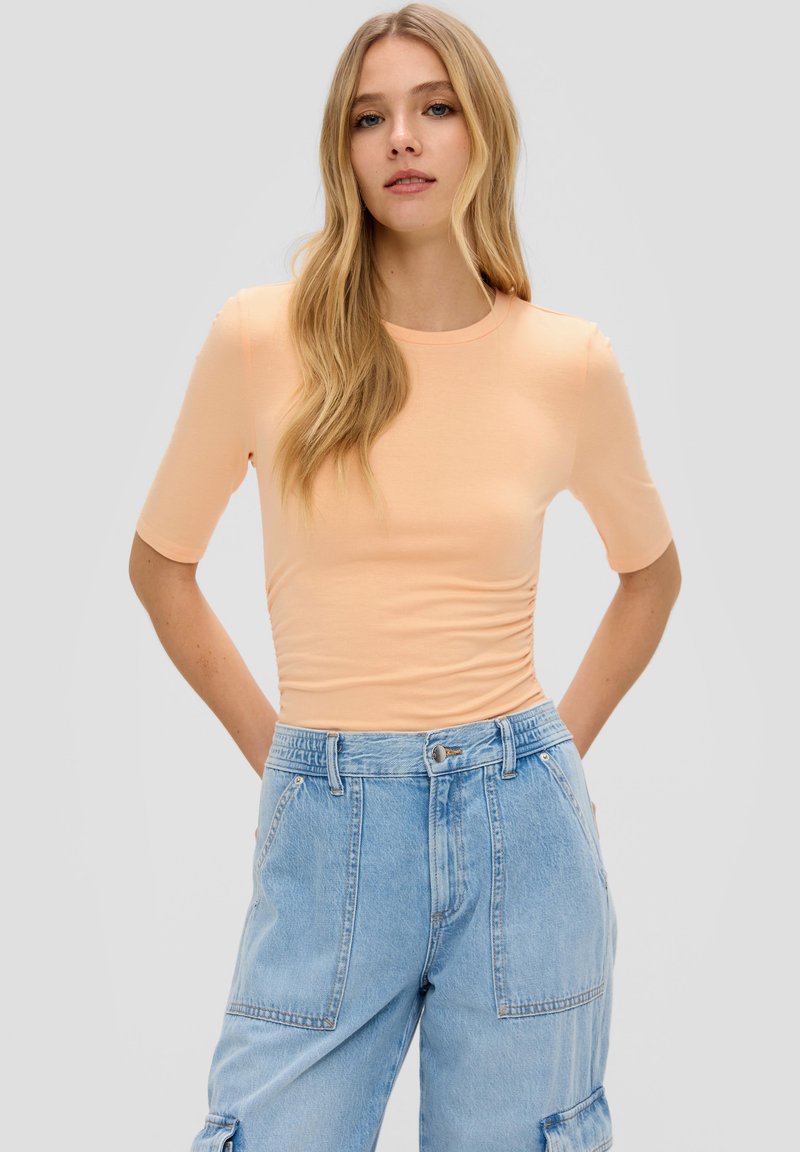 Pfirsichfarbendes, tailliertes T-Shirt mit kurzen Ärmeln, Rundhalskragen und gerafften Seitendetails, kombiniert mit hellblauen, weit geschnittenen Denim-Cargo-Hosen.