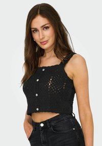 Top court en crochet noir avec un col à volants, des détails en boutons et un design texturé. Associé à un jean noir taille haute.