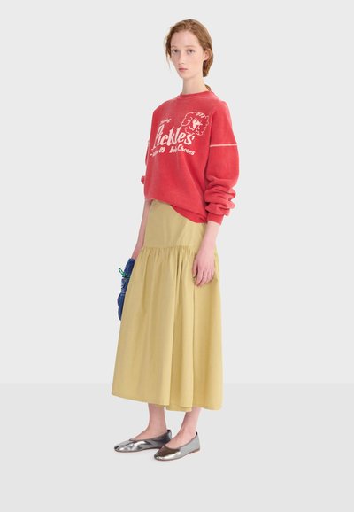 Femme portant un sweat-shirt rouge oversize avec texte, une jupe midi jaune, des ballerines argentées, tenant un sac tissé bleu, debout devant un fond uni.