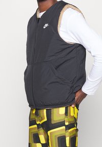 Svart vadderad väst med beige fleecekant, dragkedja fram och sidofickor, bärs över en vit långärmad skjorta och mönstrade byxor.