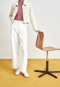 Vita vida byxor, randig röd och vit topp, krämfärgad jacka med fuskpälskrage, vita sneakers, lutande mot en trästol med metallben.