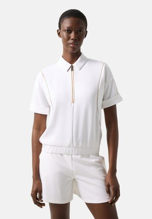 Femme portant un polo blanc à manches courtes avec fermeture éclair, coutures beige, et un short blanc assorti sur un fond uni.