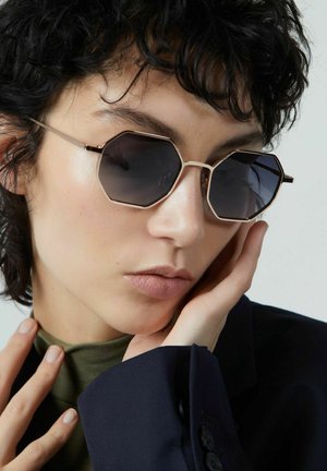 Lunettes de soleil octogonales avec des verres sombres et une fine monture en métal doré. Dotées d'une barre sourcilière proéminente et d'un subtil éclat. Portées avec un haut vert.