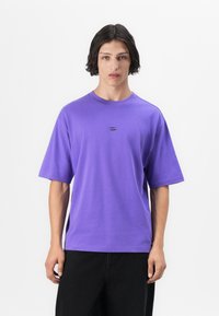 Purple short-sleeve T-shirt with a round neck, featuring a small oval emblem at the center and a relaxed fit. Made from soft cotton material.
