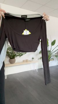 Svart langermet crop-top, rund halslinning, mykt stoff, merkelapp festet, lysegrå detaljer ved halslinningen, vist mot innendørs planter.