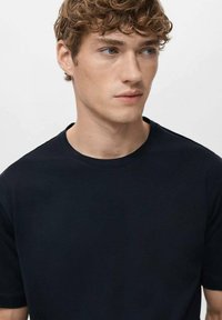 Czarna bawełniana koszulka z okrągłym dekoltem, krótkimi rękawami i gładką fakturą, noszona przez modelkę z jasnymi, kręconymi włosami.