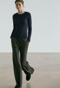 Marineblaues Langarmshirt kombiniert mit olivgrünen, weit geschnittenen Hosen mit Streifen. Bodenlang und mit glatter Textur, ergänzt durch schwarze Schuhe.