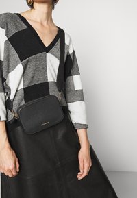 Černá kožená kabelka přes rameno se zlatými kovovými detaily a vyraženým logem. Nošena přes šedý a černý kostkovaný svetr a černou koženou sukni.