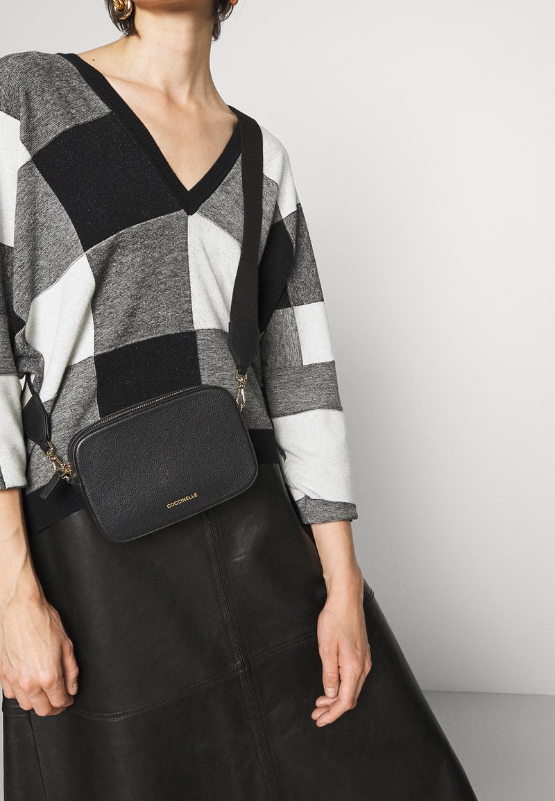 Černá kožená kabelka přes rameno se zlatými kovovými detaily a vyraženým logem. Nošena přes šedý a černý kostkovaný svetr a černou koženou sukni.