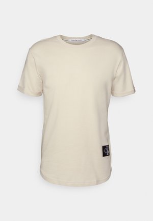 Beżowy T-shirt z krótkim rękawem i okrągłym dekoltem, z zagiętymi mankietami oraz czarną łatą z logo Calvin Klein w pobliżu dolnego brzegu.
