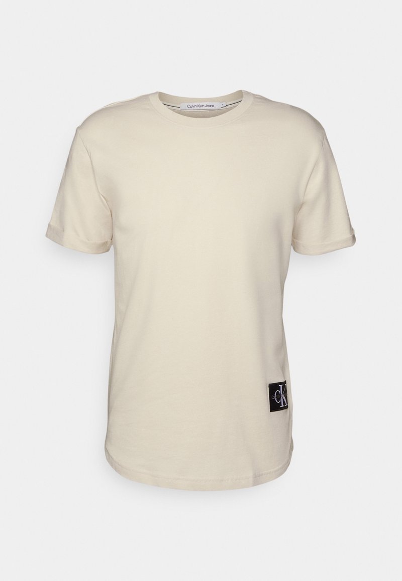 Beżowy T-shirt z krótkim rękawem i okrągłym dekoltem, z zagiętymi mankietami oraz czarną łatą z logo Calvin Klein w pobliżu dolnego brzegu.
