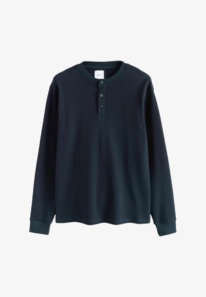 Maglietta henley a maniche lunghe in maglina waffle blu scuro con tre bottoni e polsini a coste, stesa su uno sfondo bianco.