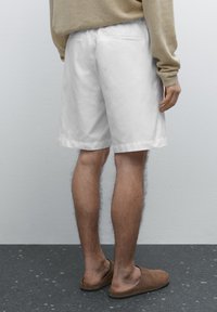 Hvide bomulds shorts med sidelommer og foldet kant, paired med brune slip-on sko. Baggrunden er en ensfarvet lys væg.