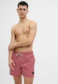 Bañador de rayas rojas con cintura ajustable, que presenta pequeños patrones en color navy y un parche de logo negro. Hecho de tejido ligero.