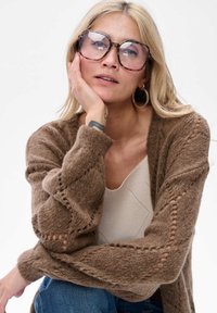 Bruine gebreide cardigan met een structuurpatroon en open mouwen, gedragen over een lichtbeige top. Grote, ronde schildpadbril en oorbellen.