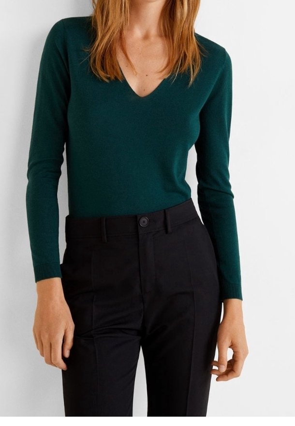 Femme portant un haut ajusté à col en V à manches longues de couleur vert foncé, rentré dans un pantalon noir taille haute, sur fond blanc.