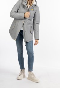 Ljusgrå puffjacka med huva, gyllene detaljer, två framfickor och emblem. Bärs över en vit skjorta och matchas med blå jeans.