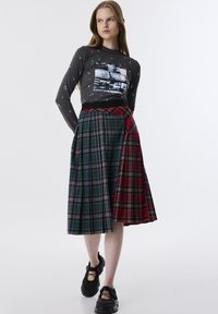 Graues Langarm-Oberteil mit Grafikdruck. Karierter Rock mit grünen, blauen und roten Abschnitten, mit Falten und kontrastierenden Texturen. Schwarze Schuhe.