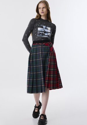 Grijze lange mouw top met grafische print. Geruite rok met groene, blauwe en rode secties, met plooien en contrasterende texturen. Zwarte schoenen.