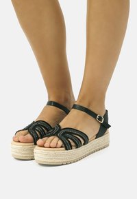 Sandali espadrilles neri con dettaglio anteriore a più strati, cinturino alla caviglia e suola in juta intrecciata. Presentano una tomaia testurizzata e un design piatto.