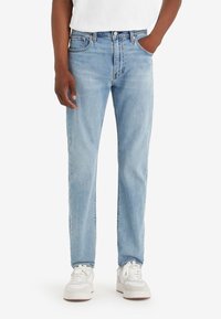 Homme portant un jean droit bleu clair, des baskets blanches et un t-shirt blanc, debout avec une main dans la poche.