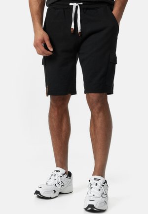 Homme portant un short cargo noir avec un cordon blanc et des baskets blanches de sport, debout devant un fond uni.