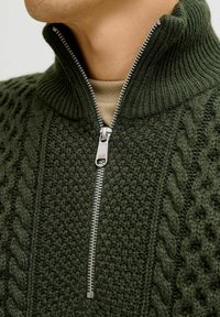 Sweater zippé vert texturé avec un col montant, présentant des motifs en tricot torsadé et un zip métallique, mettant en avant un tissu en laine épaisse.