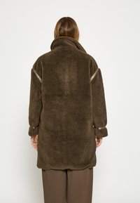 Hnedý, nadýchaný fleece kabát s vysokým golierom. Obsahuje textúrované rukávy a hladký stužkový akcent pri ramenách. Voľný strih.