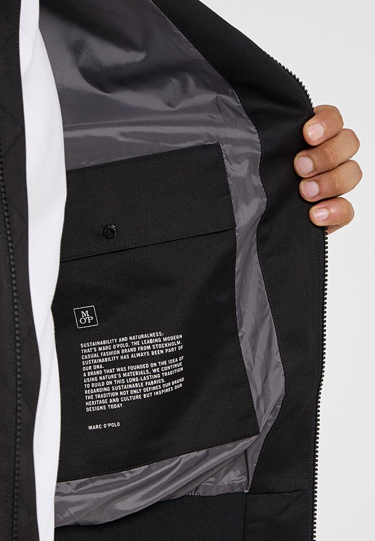 Schwarze Stoffjacke mit glattem grauem Innenfutter, einem Reißverschluss und einer Tasche mit gedruckten Markeninformationen und Logo.