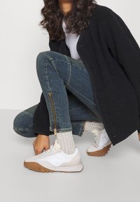 Bílé sportovní boty s texturovanou gumovou podrážkou, lehký textilní jazyk, modré džínové kalhoty s žebrovanými detaily a zipovými prvky, černý svrchní oděv.